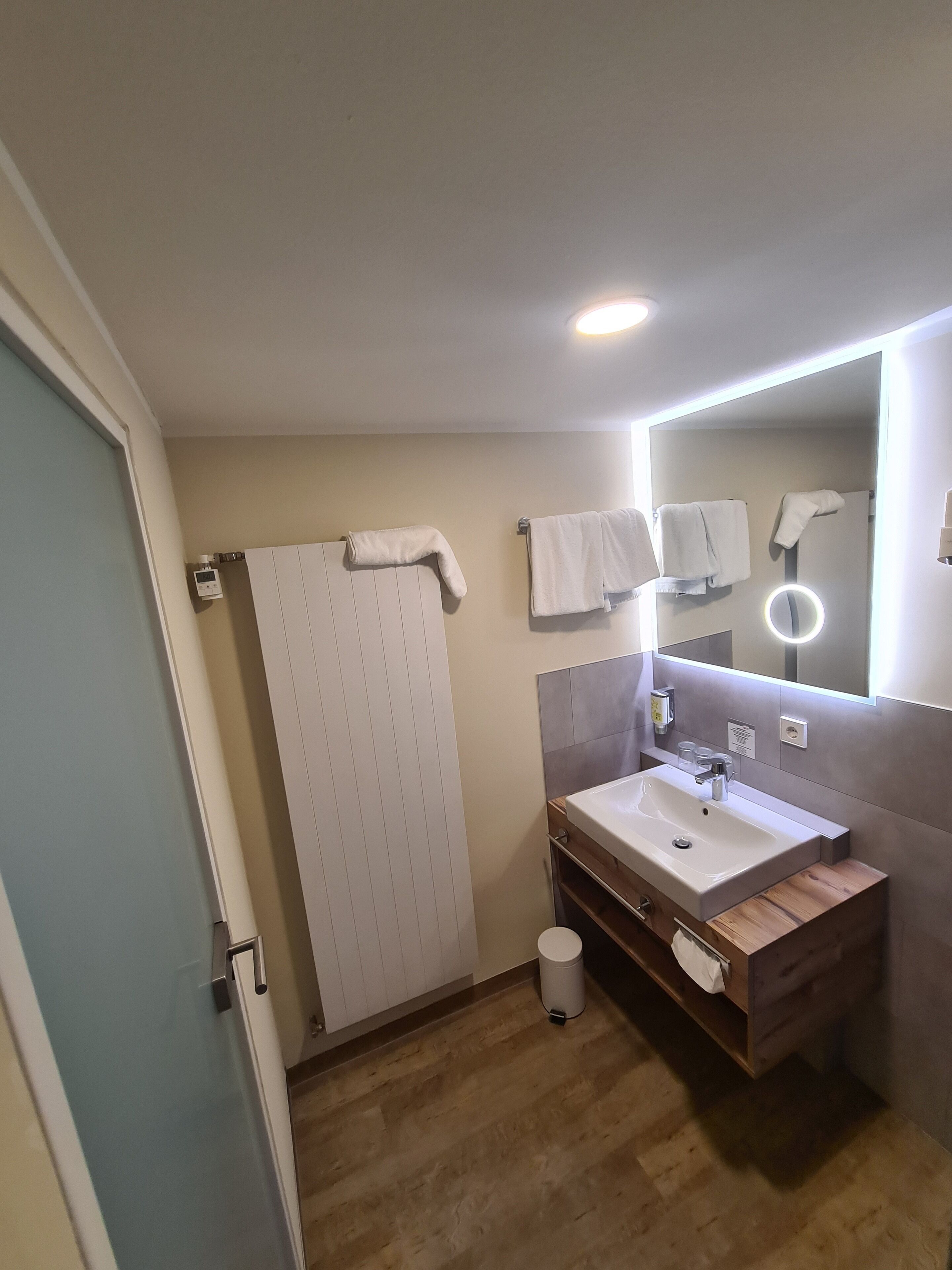 A modern bathroom with a wooden vanity, a large mirror with a ring light, and a towel rack with multiple towels.