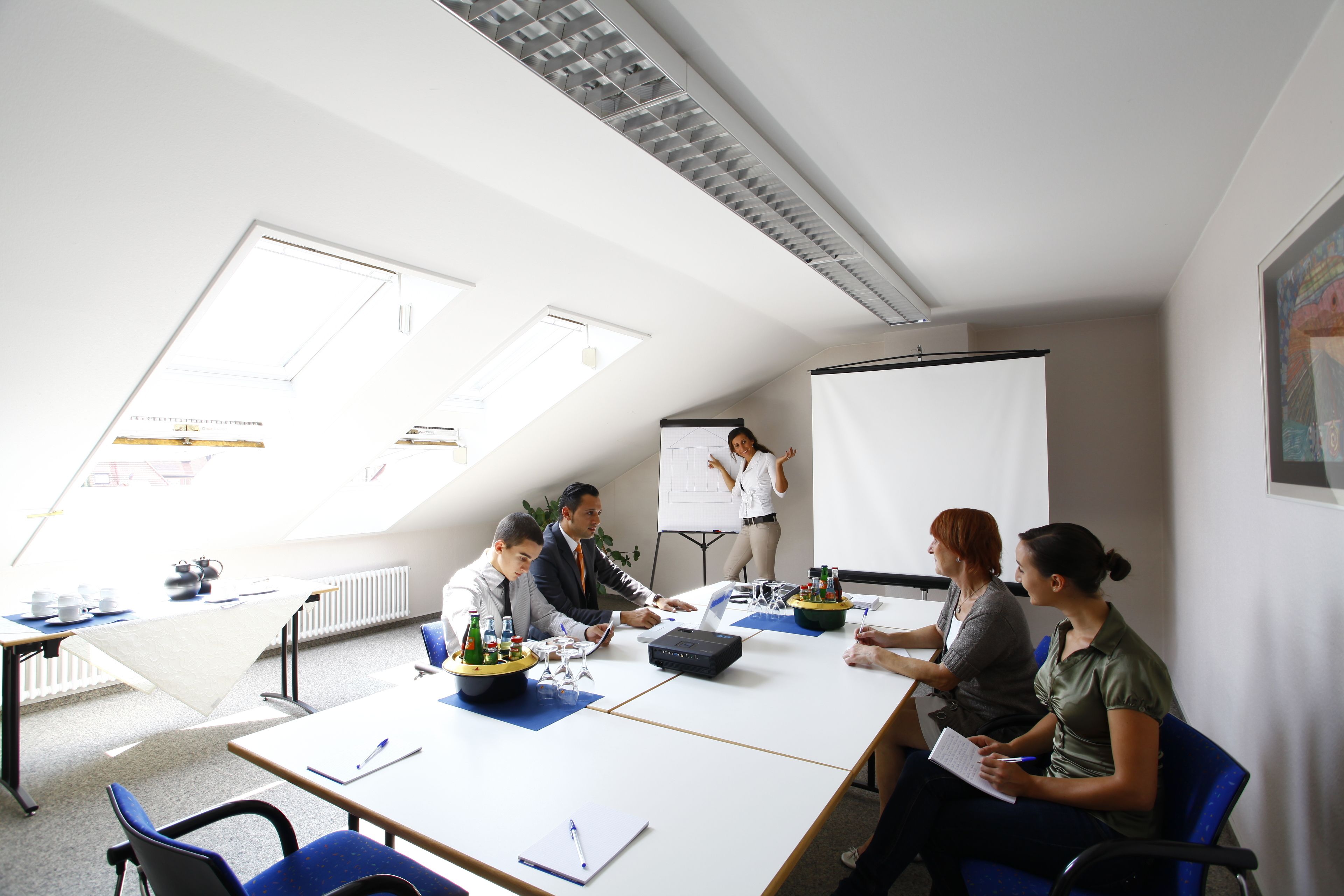 Une salle de conférence équipée d’un tableau blanc, d’un projecteur et d’un écran de présentation. On y trouve des tables avec des chaises, et une personne se tient près du tableau blanc.