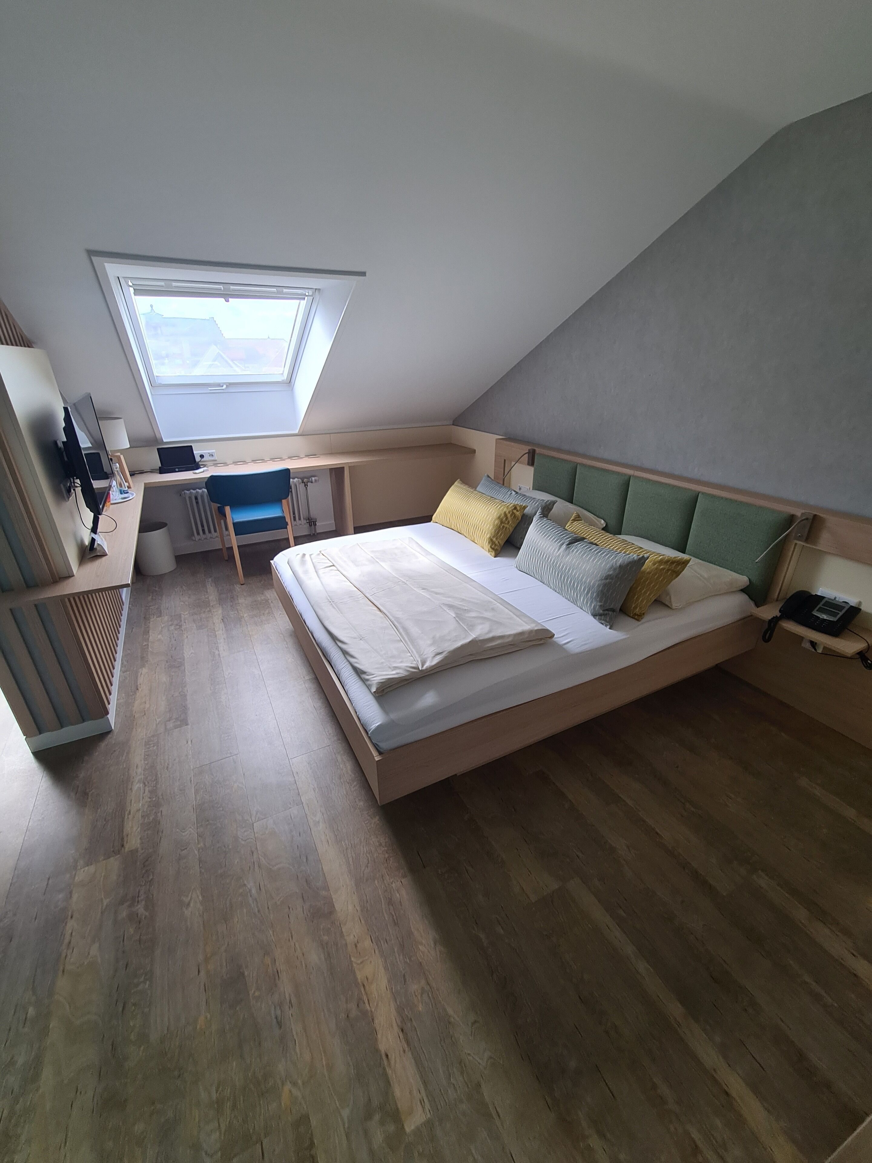 A bedroom with a sloped ceiling, a skylight, a bed with multiple pillows, a desk with a chair, and a radiator.