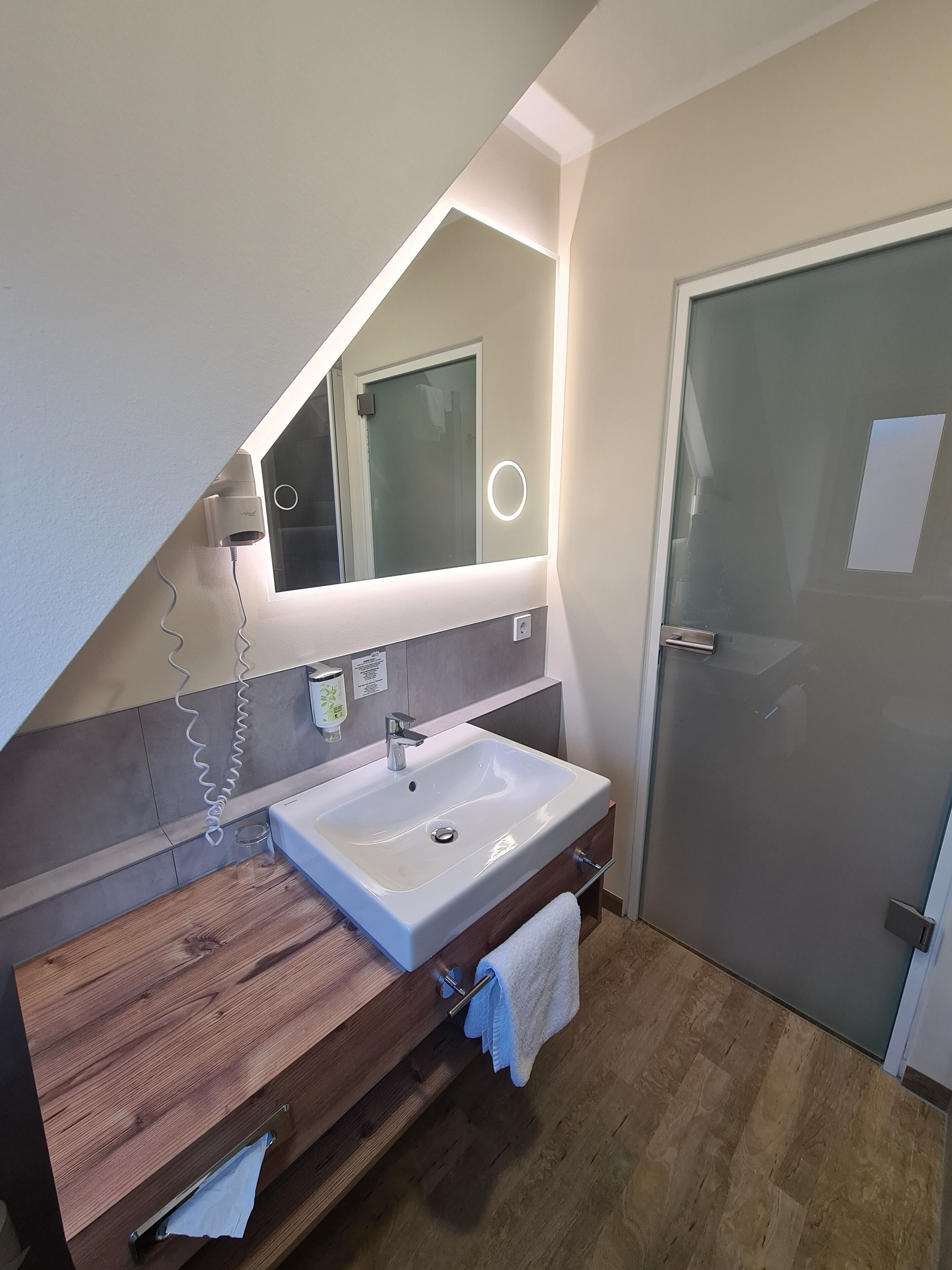Ein modernes Badezimmer mit einem weißen Waschbecken, einem Holz-Waschtisch und einem großen Spiegel mit integrierter Beleuchtung.