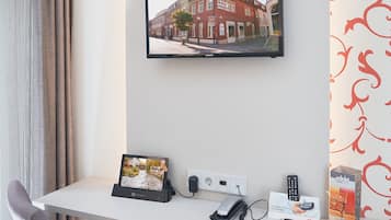 A hotel room with a desk, a chair, a flat-screen TV mounted on the wall, a remote control, a power outlet, and a small picture frame.