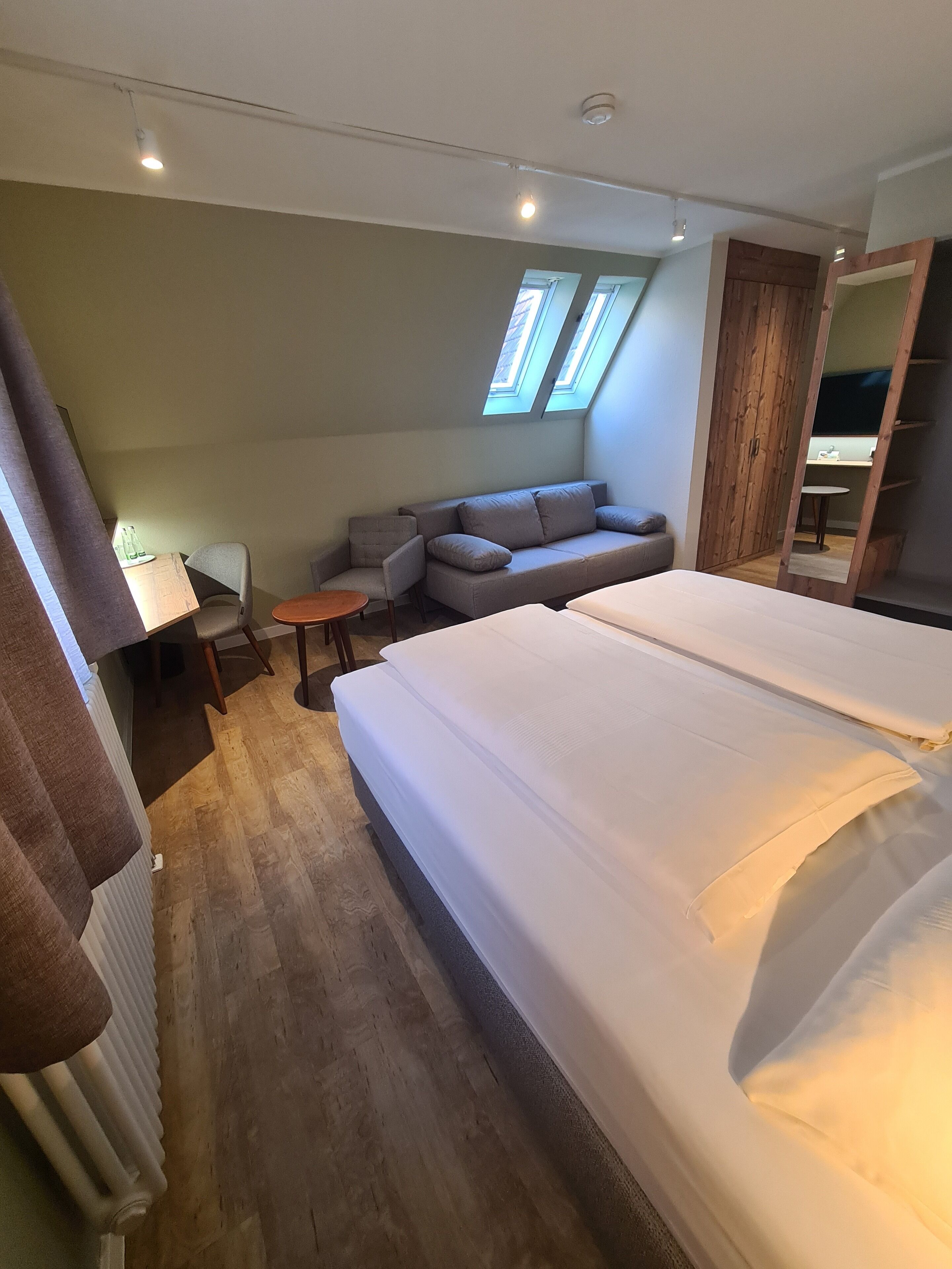 Une chambre moderne avec un grand lit, un canapé, une petite table et un radiateur. Elle est dotée de fenêtres de toit et d’une armoire.