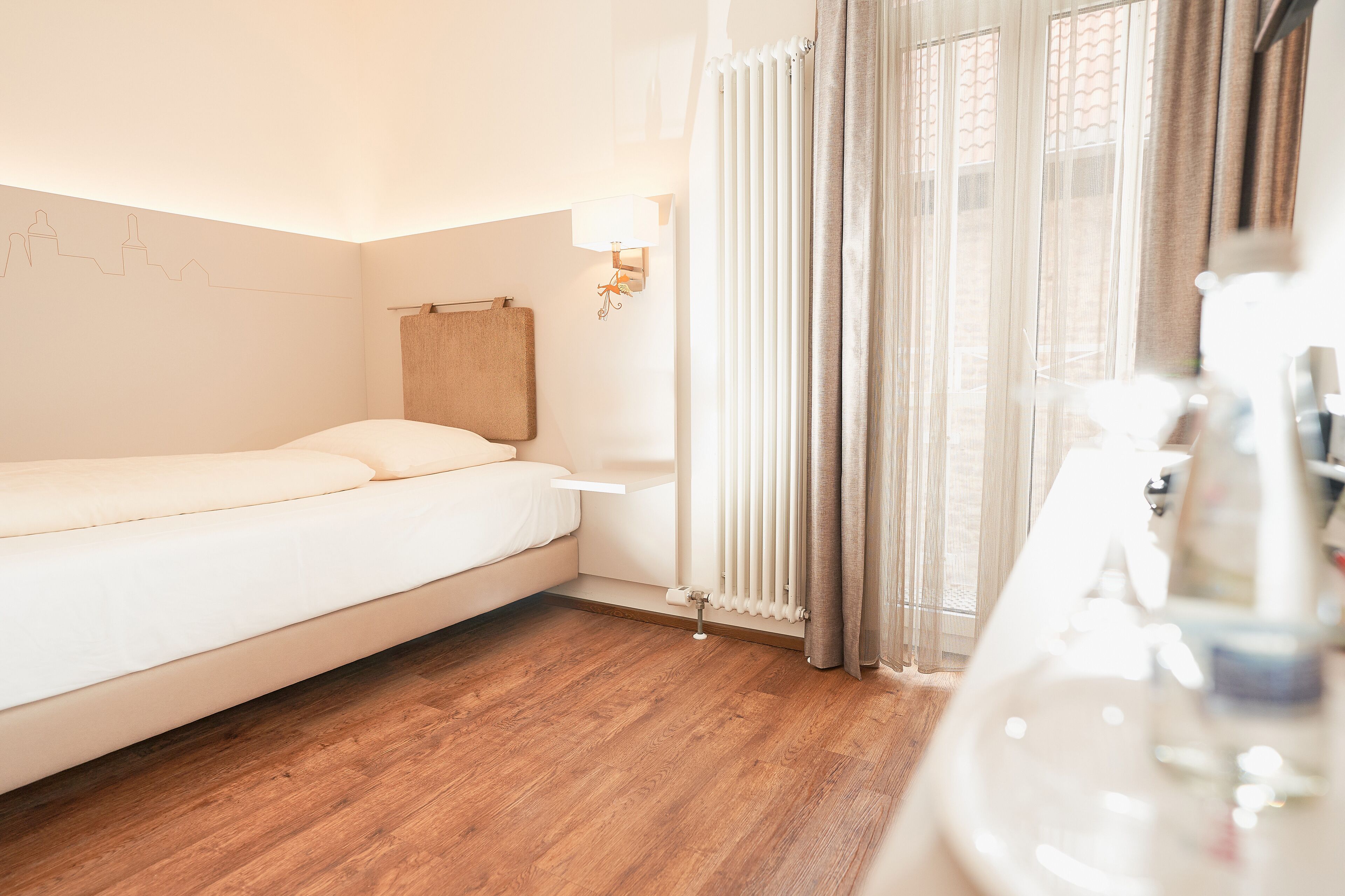 Habitación de hotel con cama, mesita de noche con lámpara, ventana con cortinas y suelo de madera.