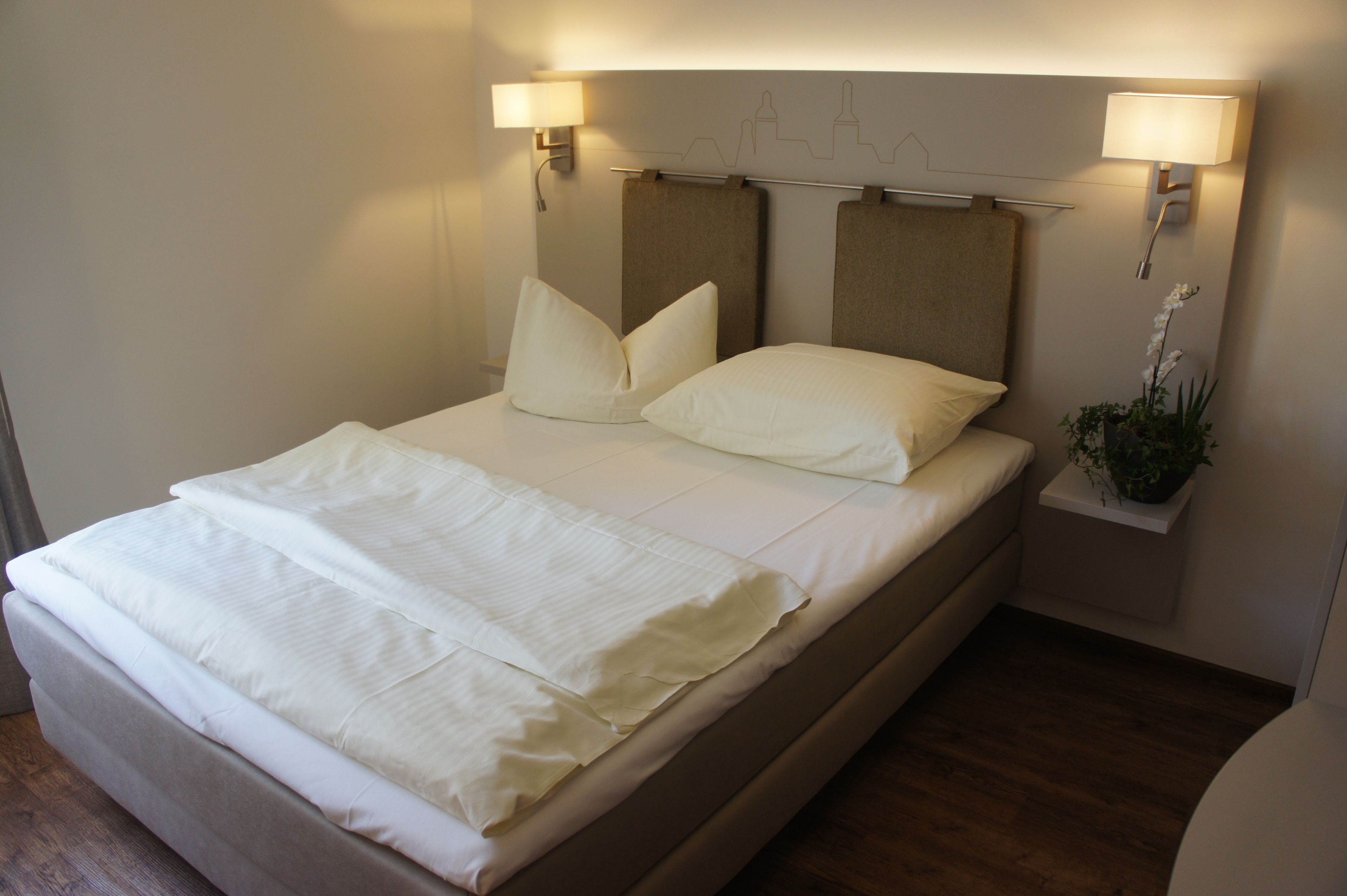 A neatly made bed with white linens, two wall-mounted lamps, and a small plant on a nightstand.