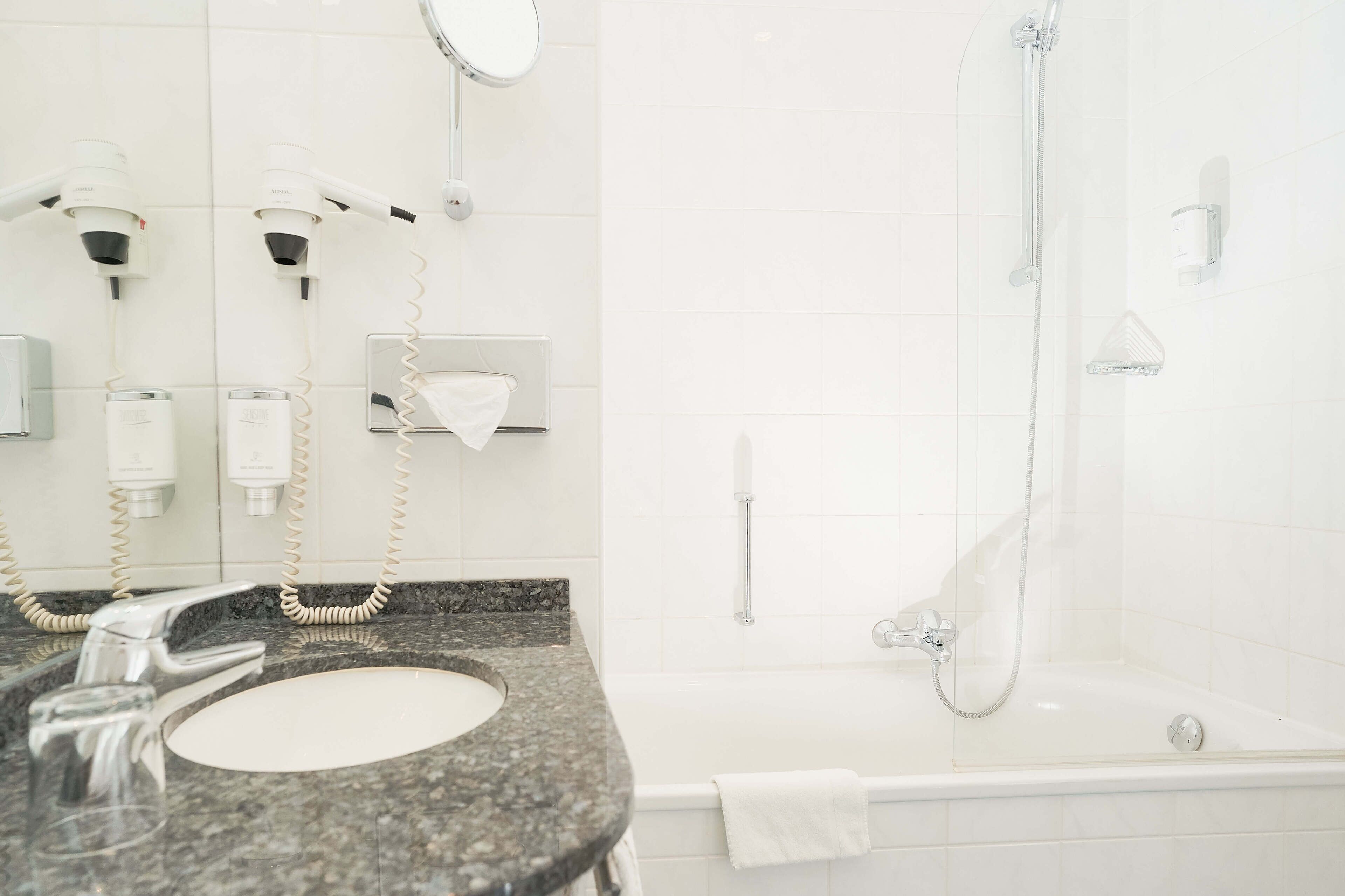 Een badkamer met een wastafel met granieten blad, een spiegel, een handdoekhouder, een handdouche en een bad.