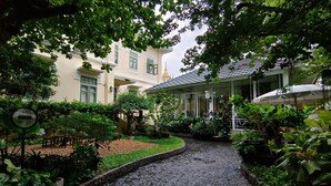 A cobblestone pathway in a lush garden area with potted plants and a building with a tiled roof. - Baan2459 Heritage House (Bangkok)