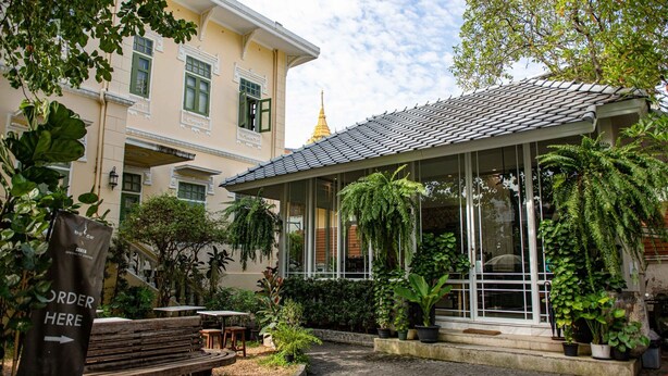 A building with a covered patio area, potted plants, and a sign indicating open hours. - Baan2459 Heritage House (Bangkok)