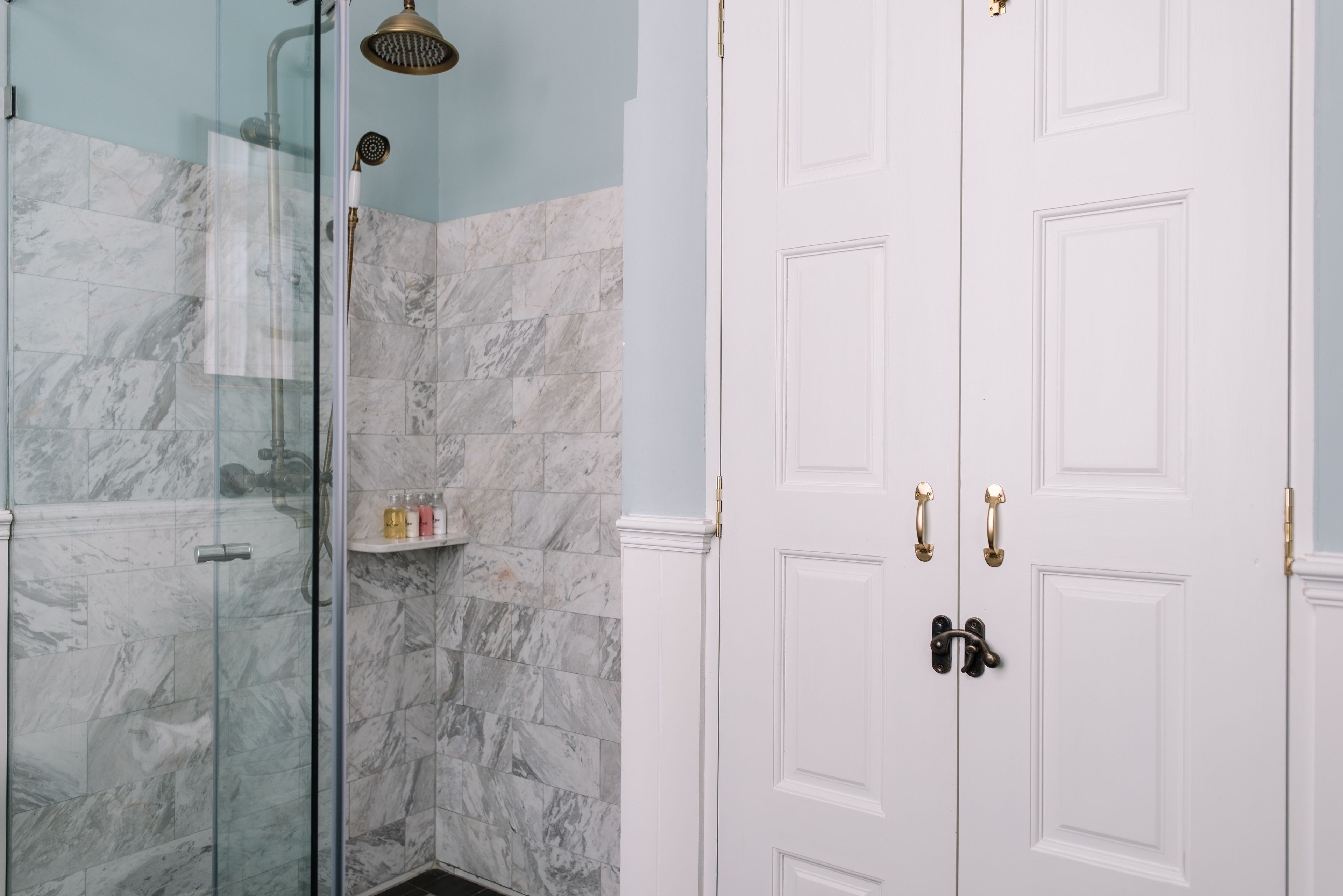 Une salle de bain dotée d’une douche à parois de verre, de murs en marbre et d’une porte blanche avec des poignées en laiton.