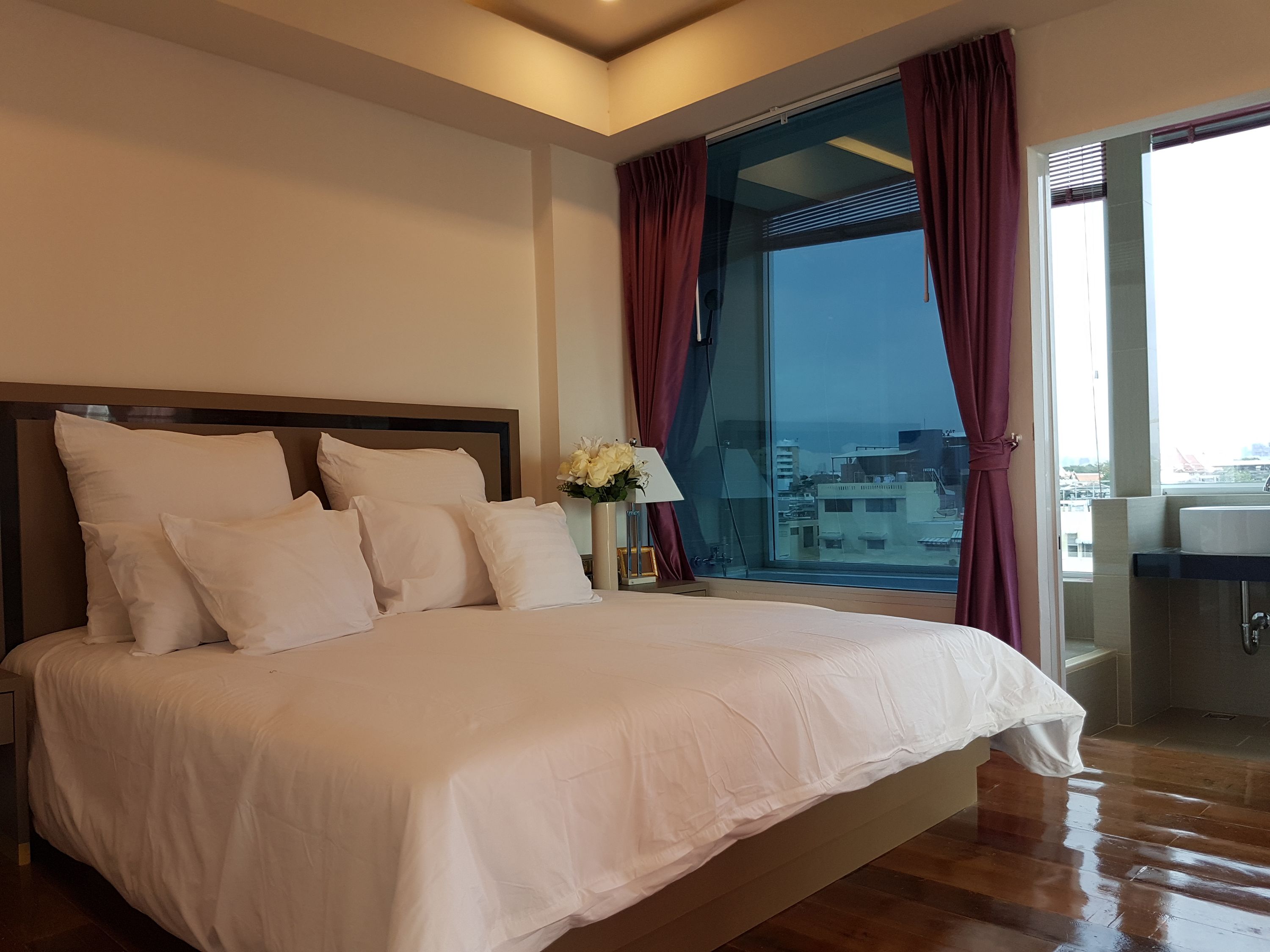 A bedroom with a large bed, a nightstand with a lamp, and a view of the city through a large window.