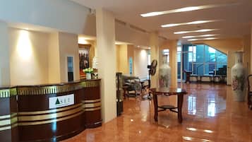 A hotel reception area with a reception desk, chairs, a vase, and a staircase in the background.