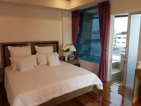 Habitación de hotel con una cama grande, una mesita de noche con lámpara, un florero con flores y vistas a la ciudad a través de una ventana con cortinas.