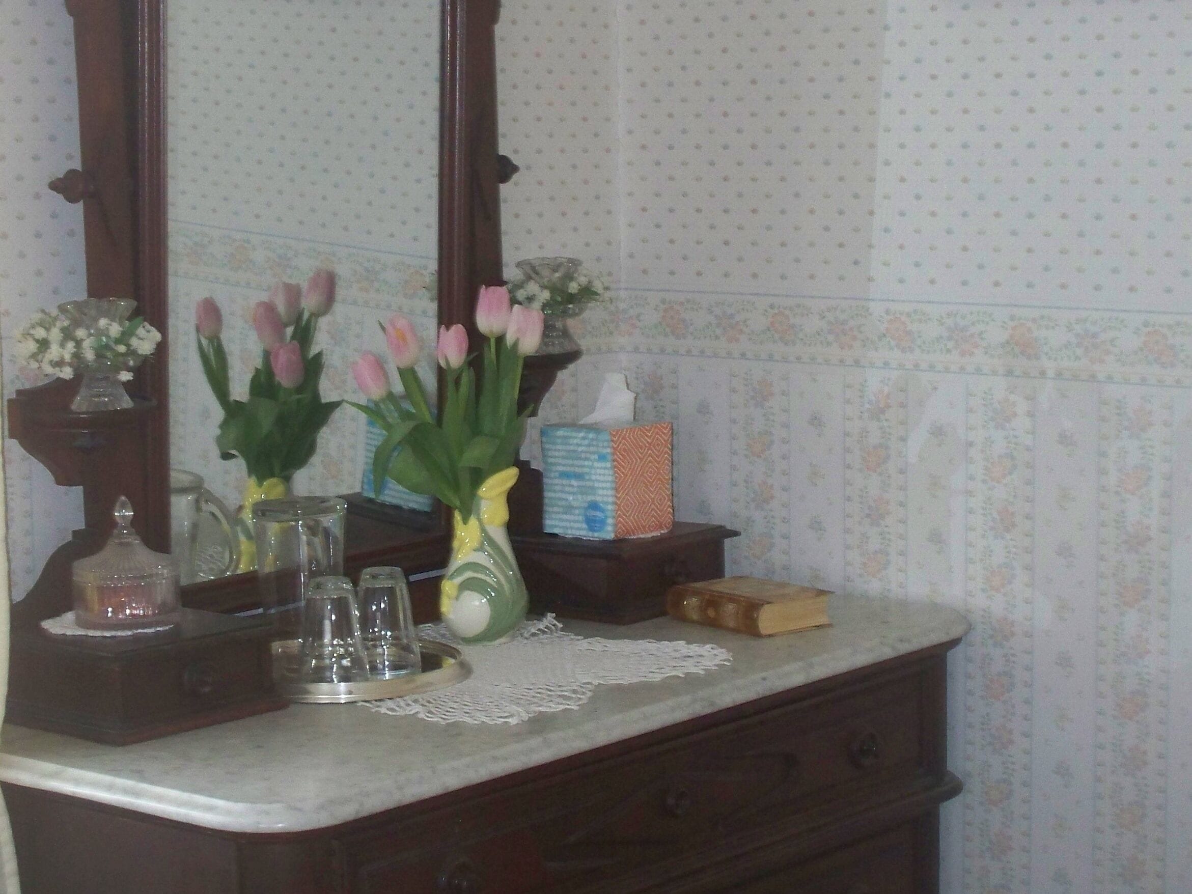 A bathroom vanity with a mirror, a vase with pink flowers, a tissue box, and a glass pitcher.