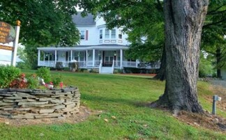 A large white house with a porch, surrounded by trees and a stone wall.