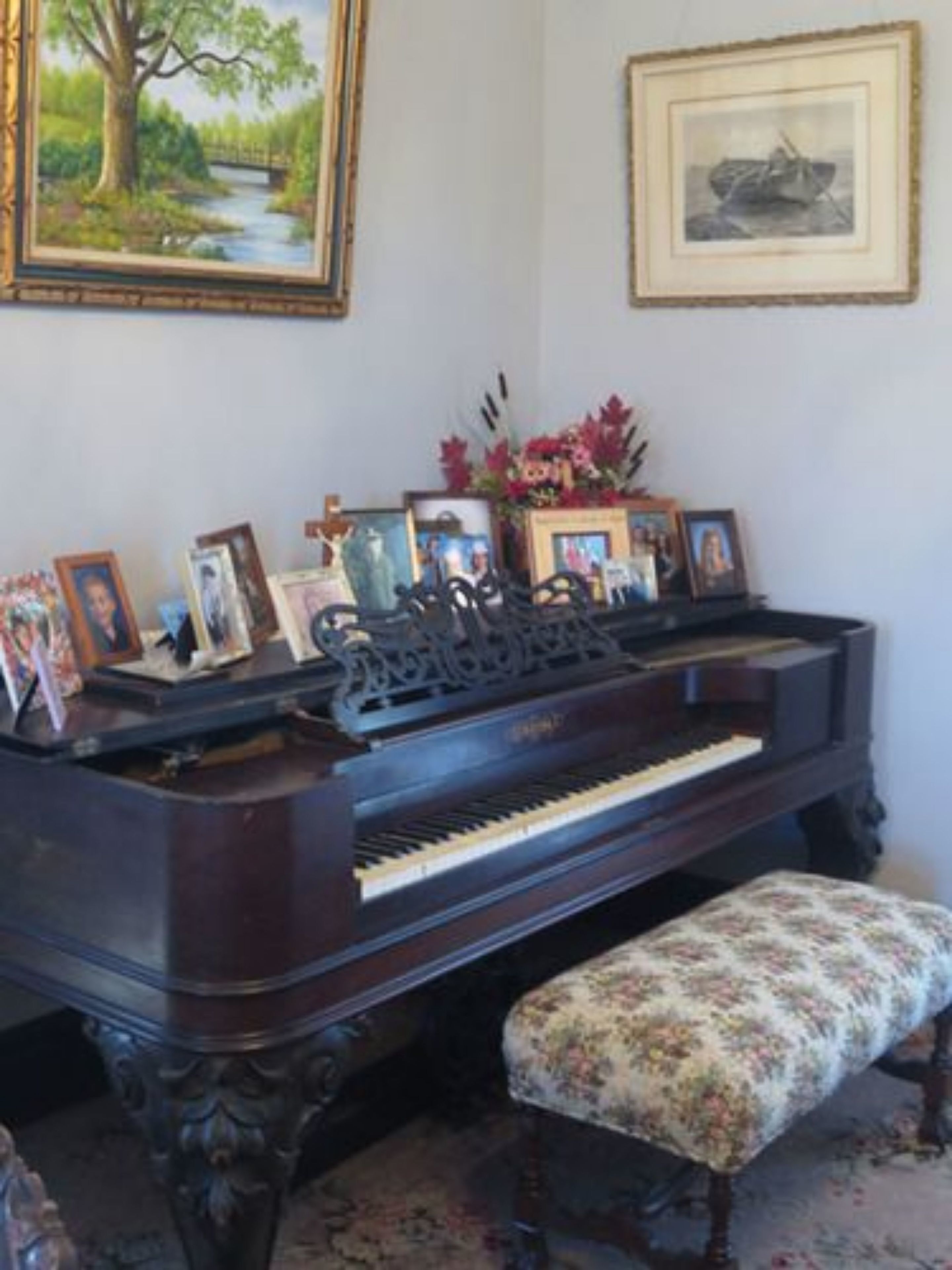 Un piano à queue avec un banc, entouré de tableaux encadrés et d’un vase de fleurs.