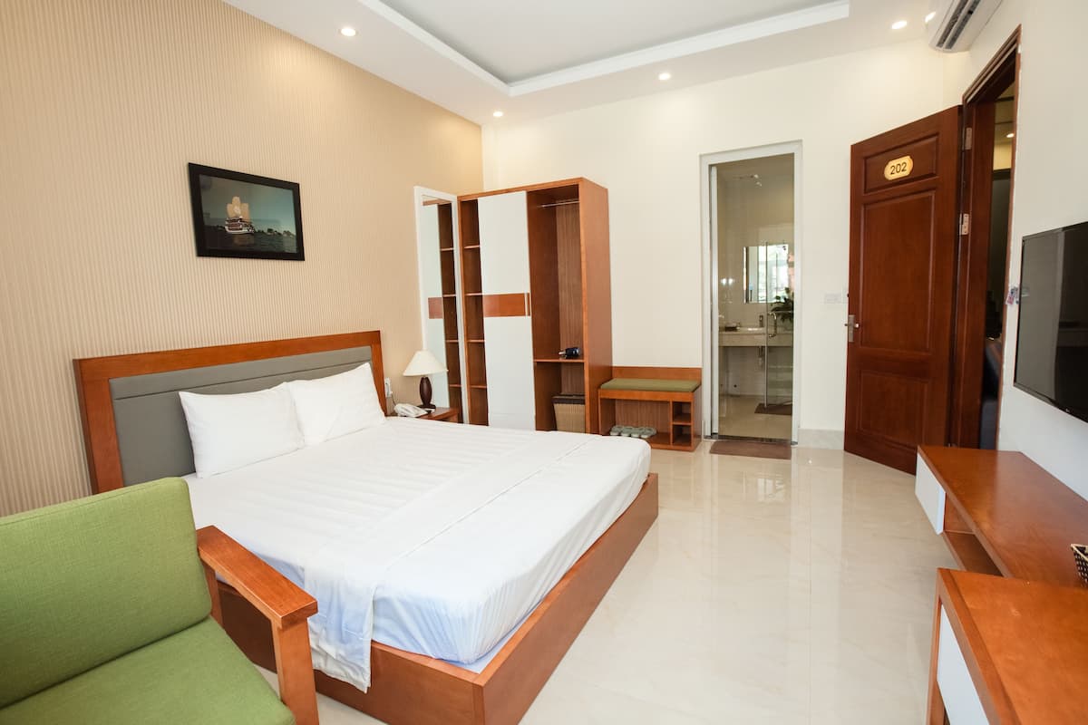 A hotel room with a large bed, a green armchair, a wooden wardrobe, a television, and a bathroom visible through an open door.