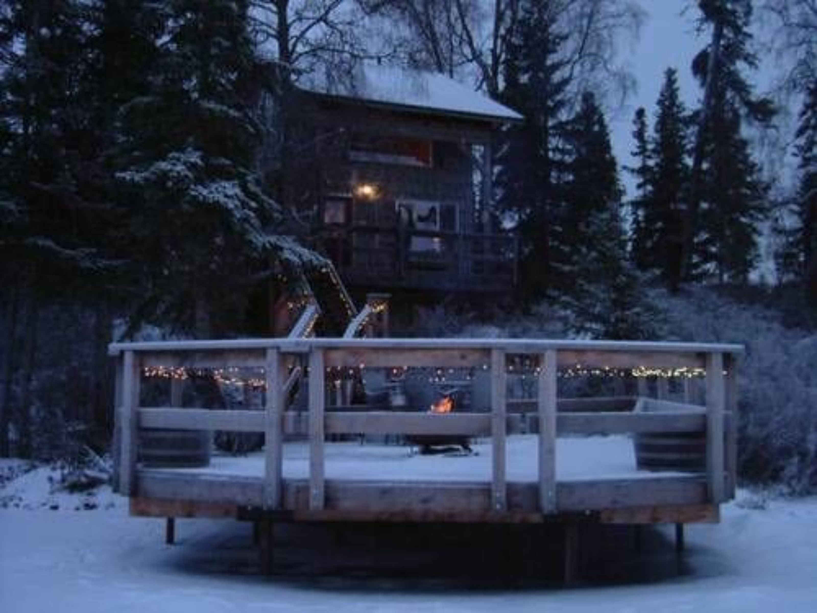 Une terrasse en bois avec une rambarde, entourée d’arbres couverts de neige et d’un bâtiment en arrière-plan.