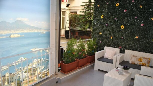 Un balcón con muebles de mimbre blanco, plantas en macetas y vistas a un puerto deportivo y al paisaje urbano.