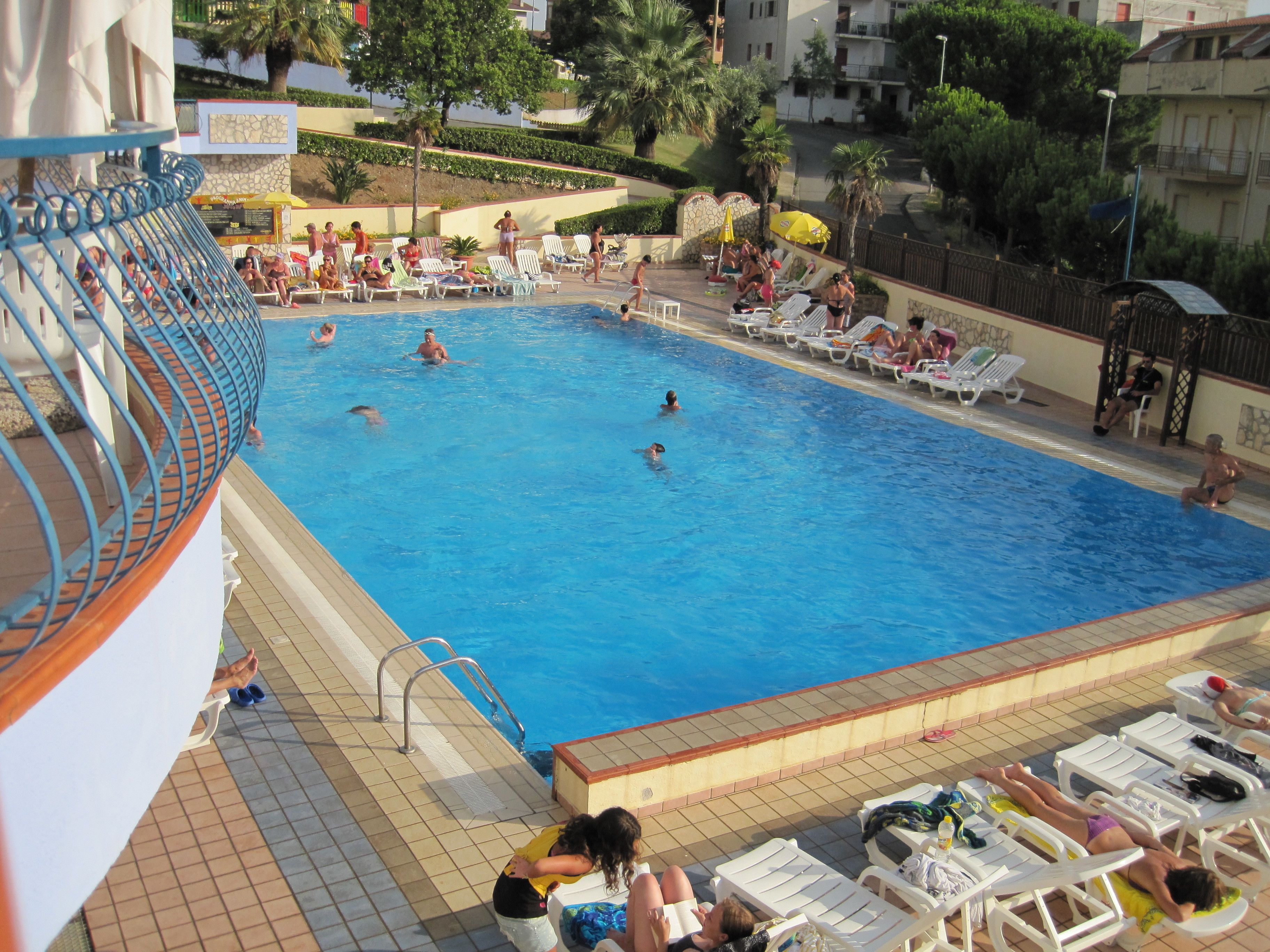Une piscine où des gens nagent et prennent des bains de soleil. Des chaises longues et des parasols sont disposés autour de la piscine.