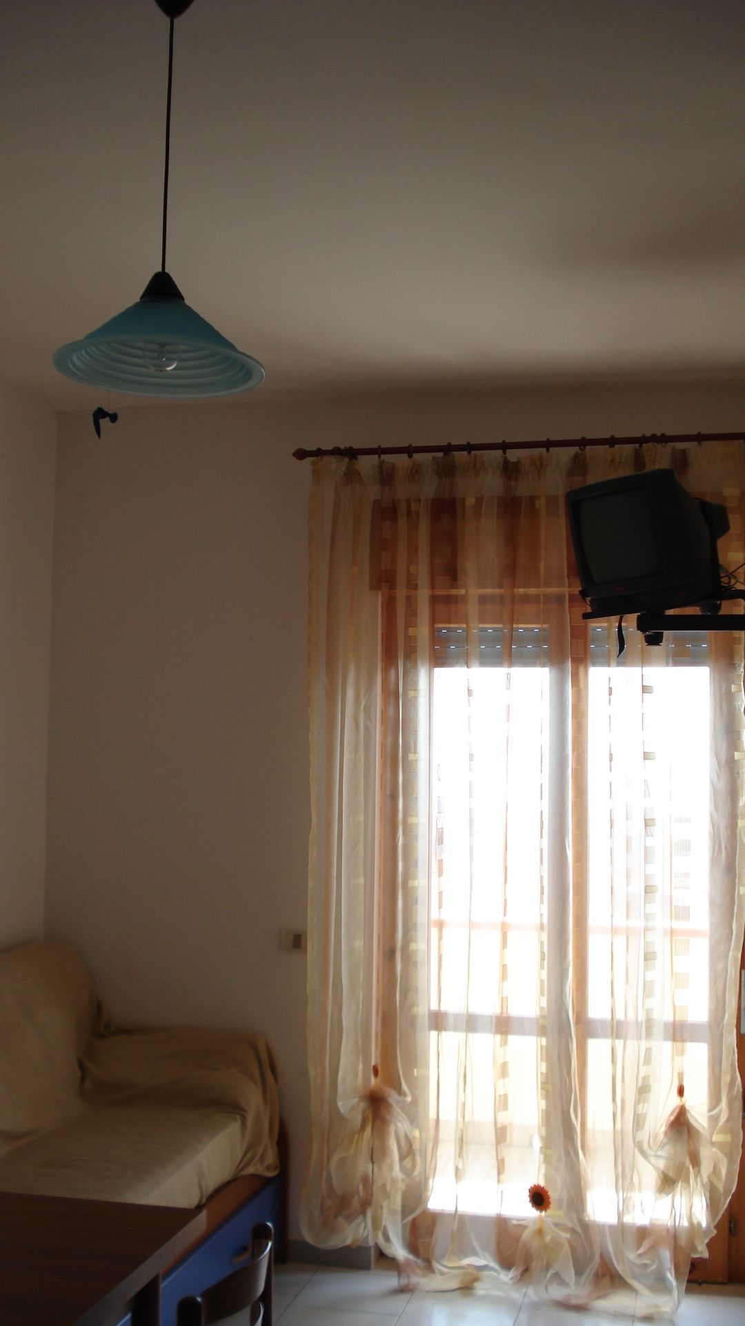 A room with a bed, a television mounted on the wall, and a window with sheer curtains.