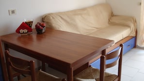 A small, cozy room with a wooden table, two chairs, a sofa, and a small table with a glass, fruits, and a red cloth.