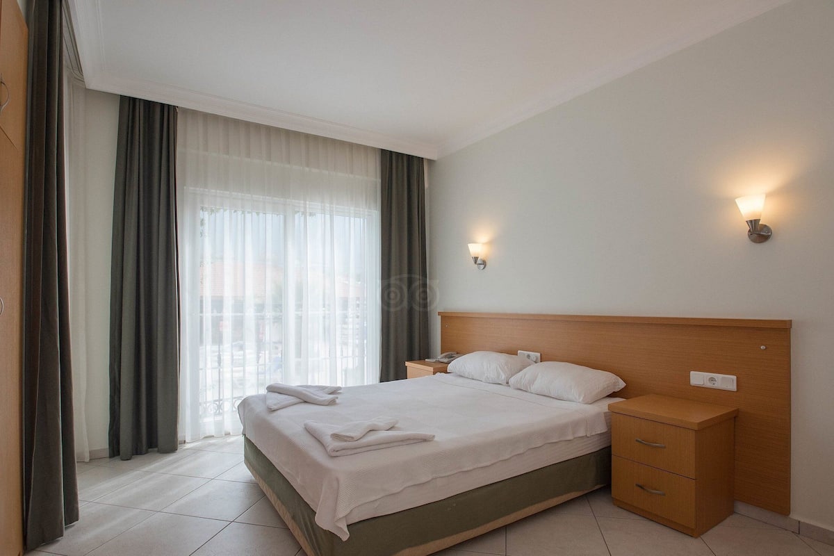 Una habitación de hotel con una cama, mesitas de noche, una lámpara y una ventana con cortinas.