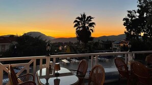 Una terraza con sillones de mimbre y una mesa, con vista a un atardecer y montañas al fondo.
