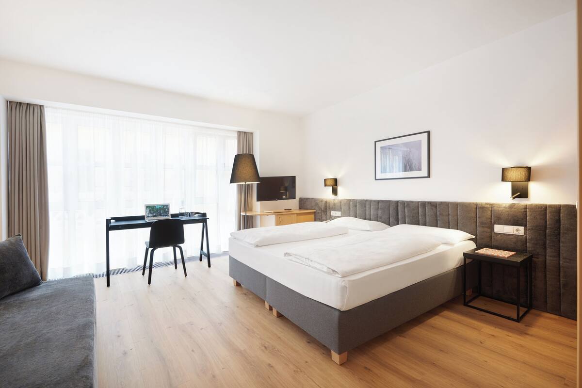 A modern hotel room with a large bed, a desk, a chair, and a sofa.
