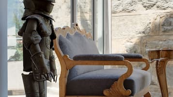 A statue of a knight in armor next to a wooden chair with a cushion, in a room with stone walls and a large window.
