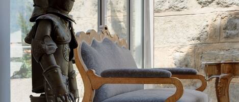 A statue of a knight in armor next to a wooden chair with a cushion, in a room with stone walls and a large window.
