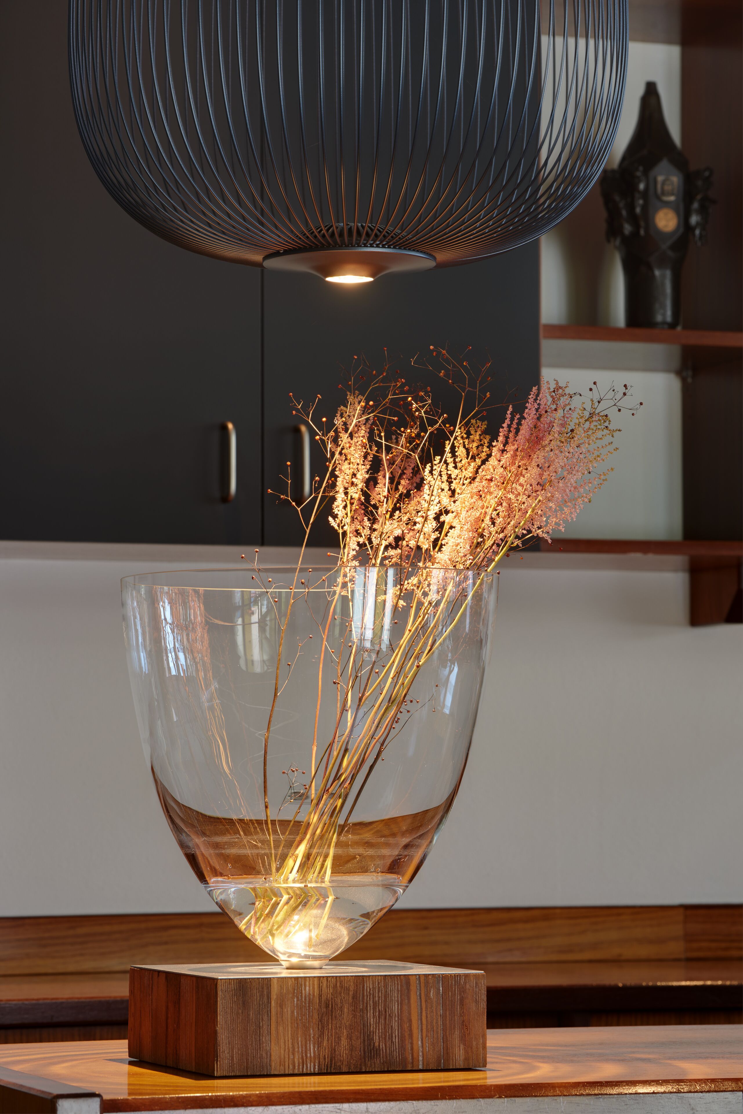 A clear glass vase with dried flowers on a wooden base, a dark-colored cabinet in the background, and a modern pendant light above.