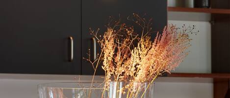 A clear glass vase with dried flowers on a wooden base, a dark-colored cabinet in the background, and a modern pendant light above.