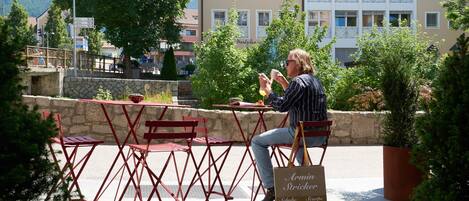 A person sitting at a table with a red chair, holding a cup and a smartphone. There is a sign with text on it, and a potted plant nearby.