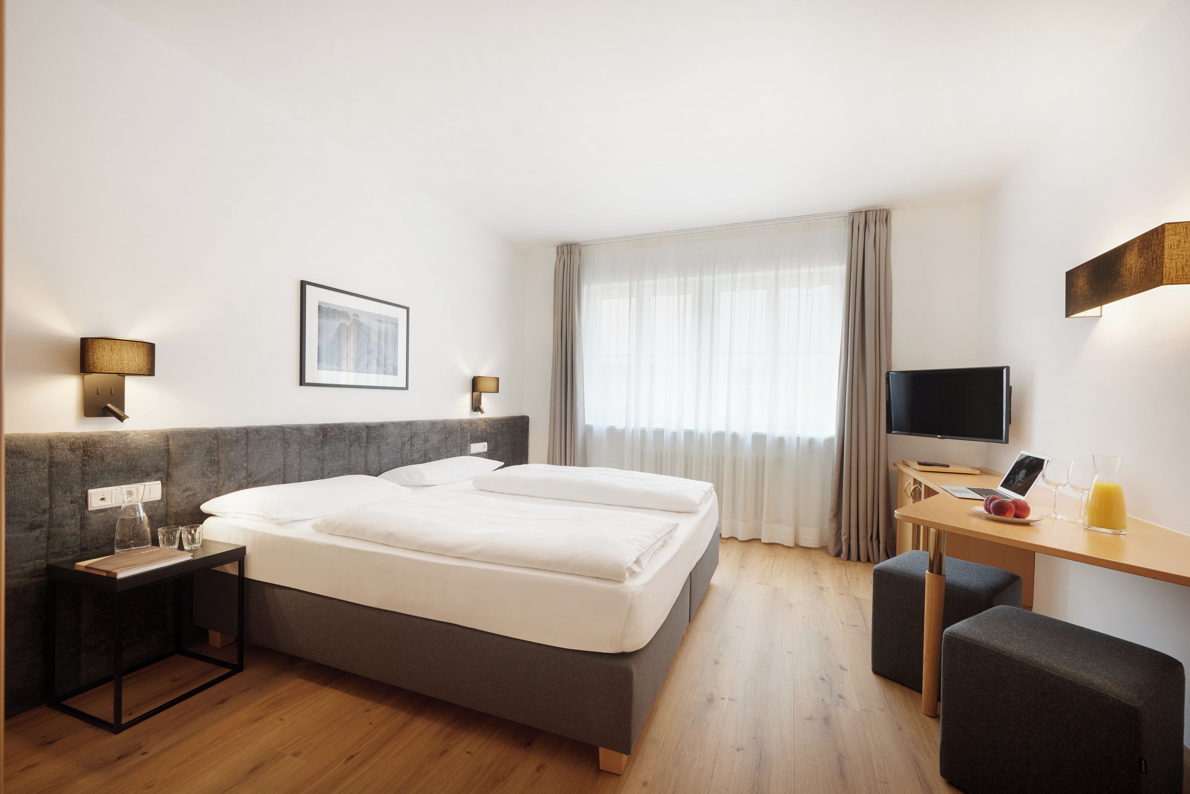 Habitación de hotel con cama, mesitas de noche, escritorio con silla, televisor y ventana con cortinas.