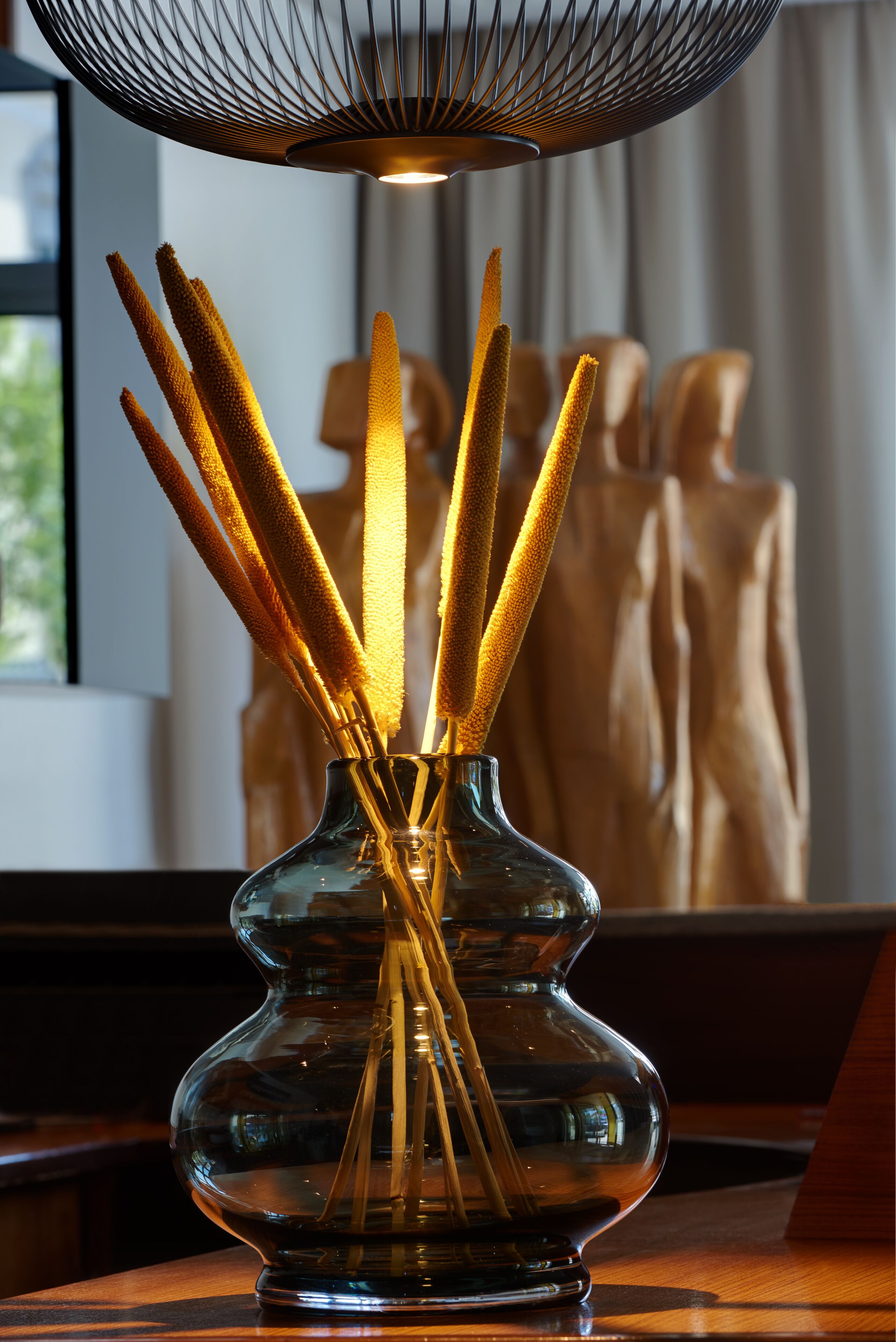 A clear glass vase with reed sticks on a wooden surface, a hanging wire light fixture above, and a blurred background with a window and a mannequin.