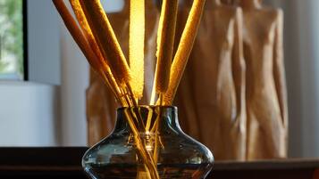 A clear glass vase with reed sticks on a wooden surface, a hanging wire light fixture above, and a blurred background with a window and a mannequin.