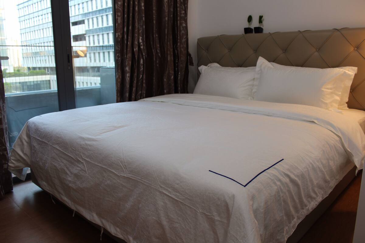 A hotel room with a large bed, a tufted headboard, and a view of a cityscape through a large window.