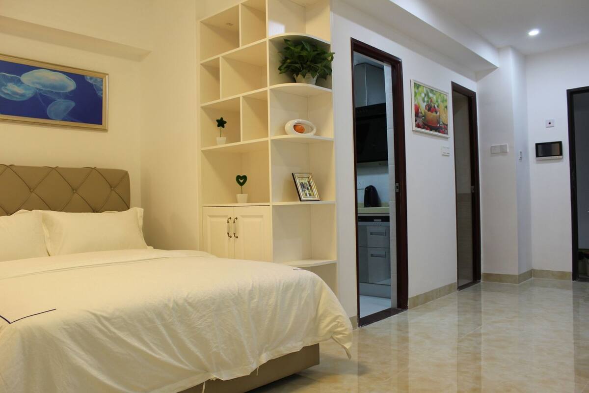 A bedroom with a bed, a white wardrobe, a plant, and a painting on the wall.