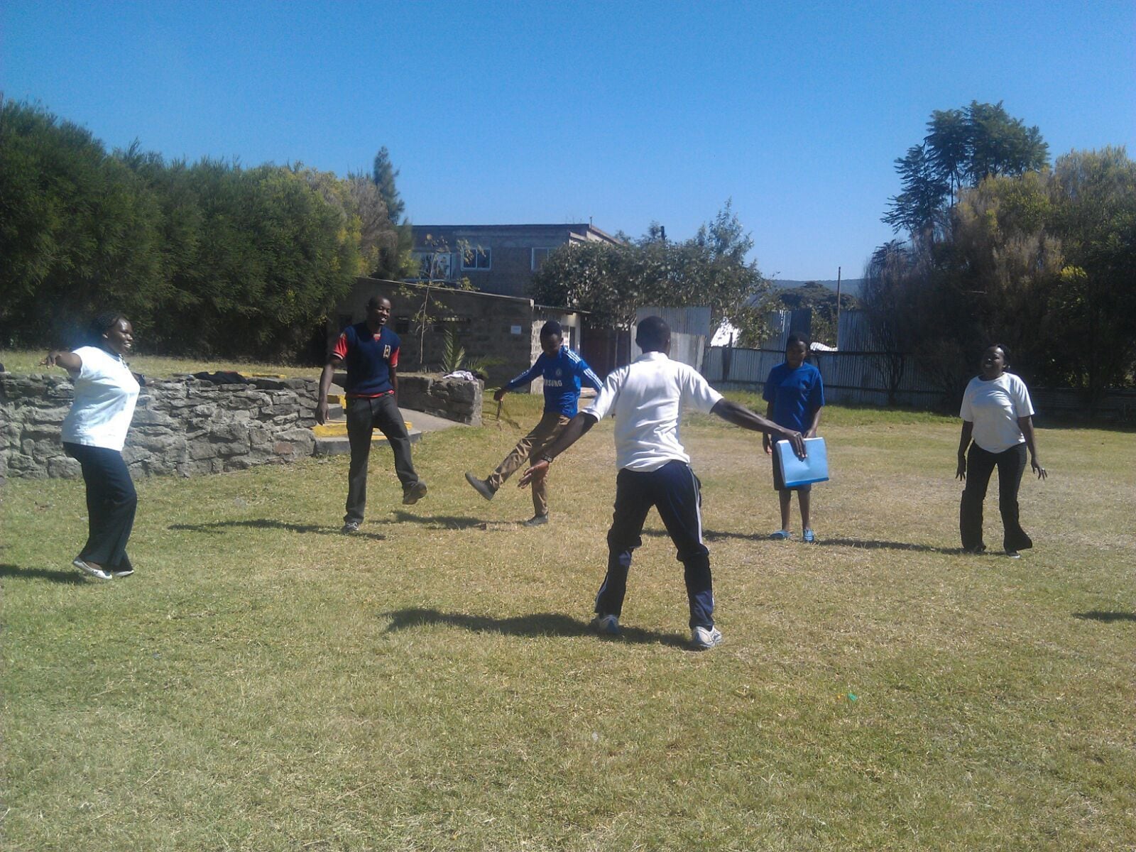 A group of people playing a sport in an outdoor setting with a stone wall and trees in the background.
