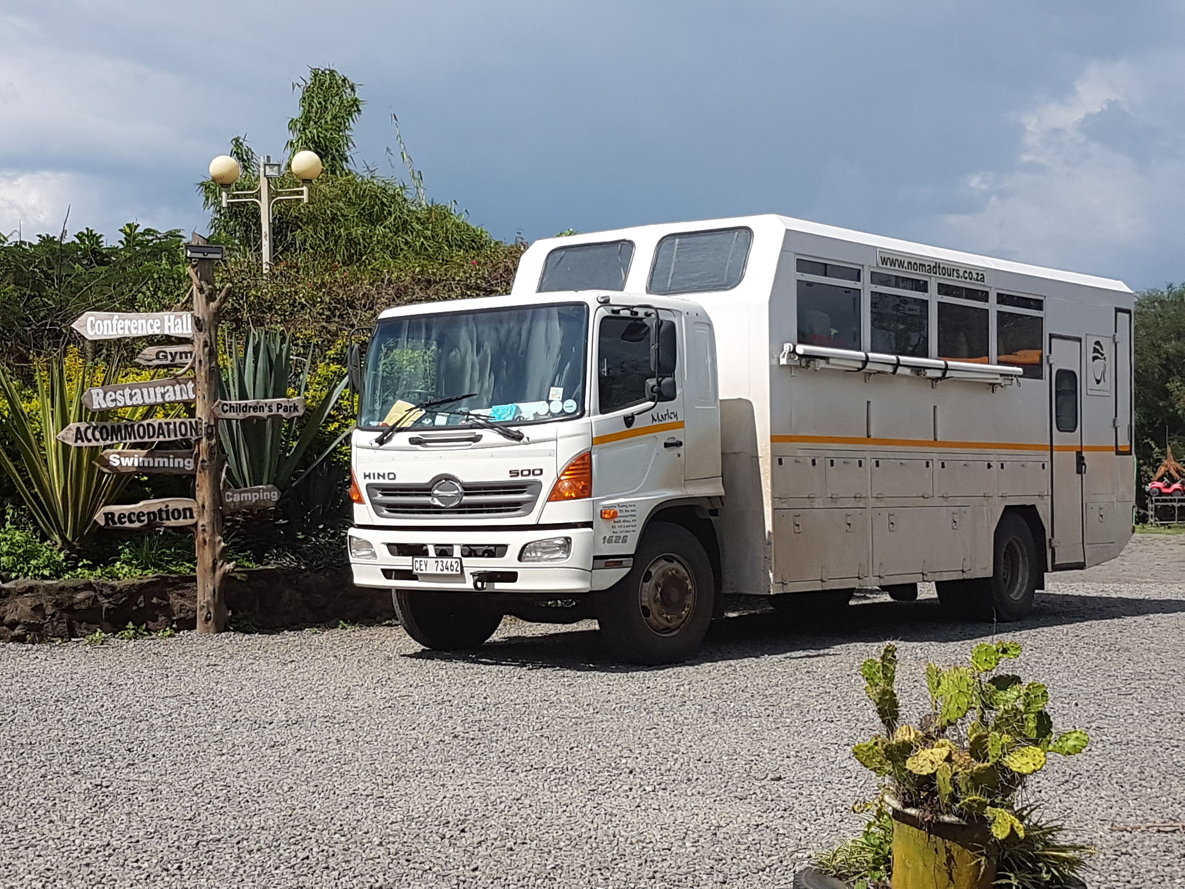 Uma caminhonete Hino 500 branca estacionada em superfície de cascalho com um poste de sinalização listando comodidades como salão de conferências, academia, restaurante e piscina.