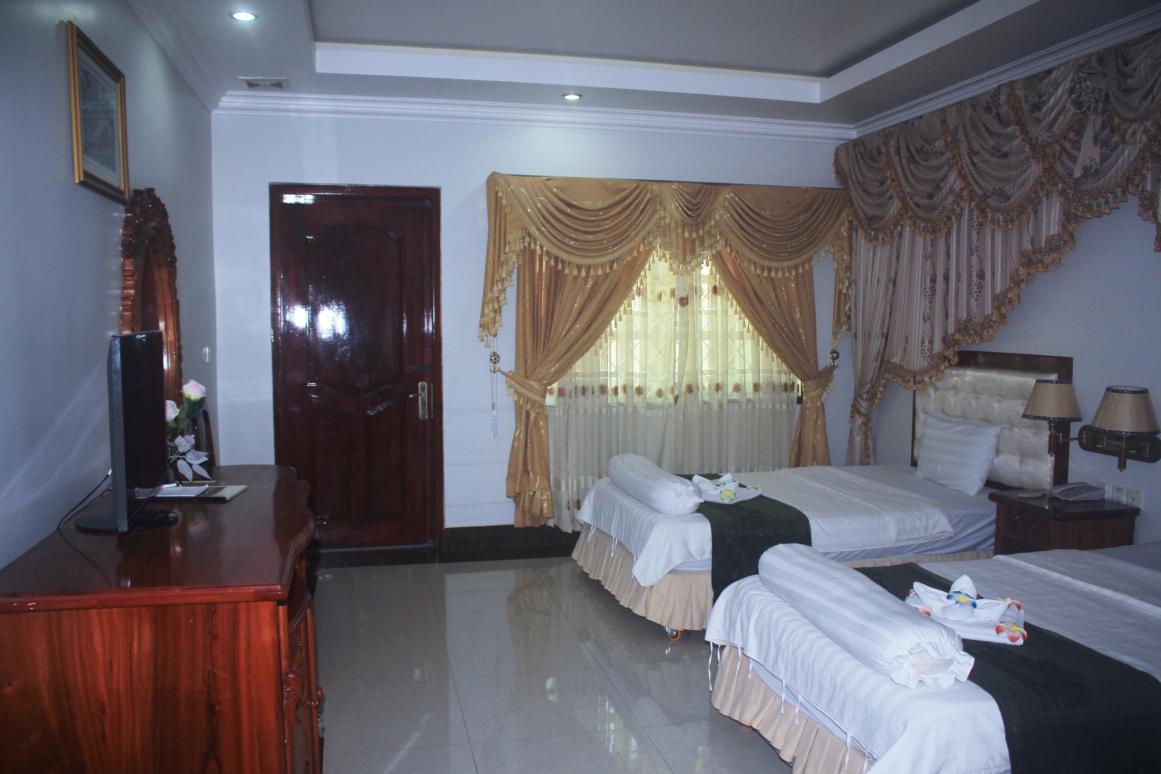 A hotel room with two beds, a wooden desk, a television, and a large window with curtains.
