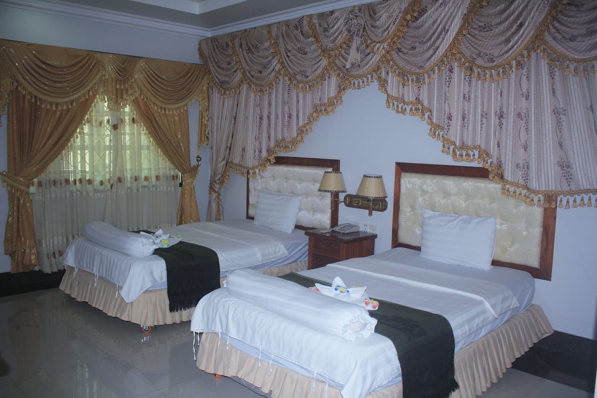 A hotel room with two beds, each with a headboard, a nightstand with lamps, and a window with curtains.