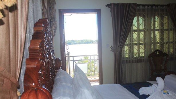A hotel room with a bed, a wooden headboard, a window with a view of a body of water, and a balcony. - Monorom 2 VIP Hotel (Kampong Cham)