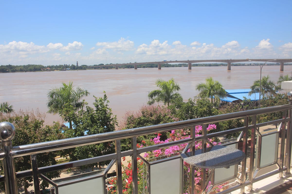 A view of a river with a bridge in the background, surrounded by greenery and pink flowers.