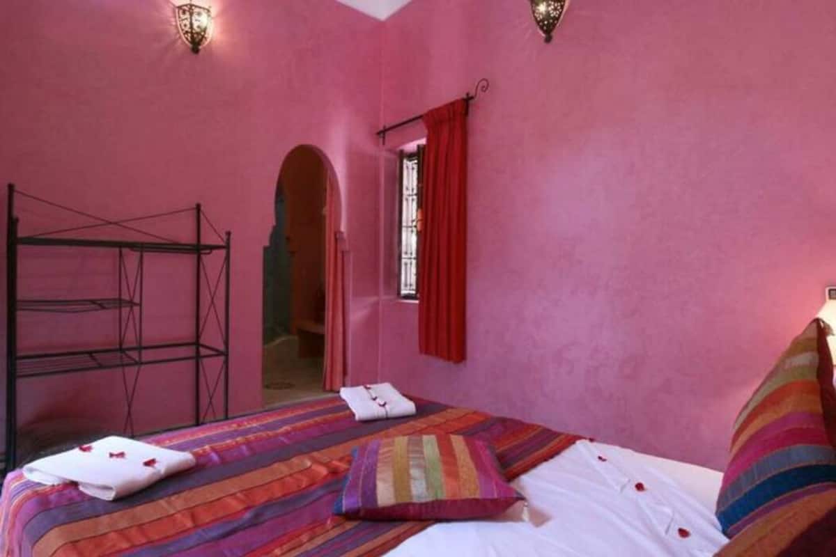 A pink-themed room with a bed, a window with red curtains, and a metal rack.