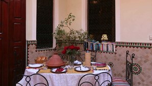 A dining area with a table set for two, a Moroccan tagine, and a bookshelf with books. - Riad Todra (Marrakech)