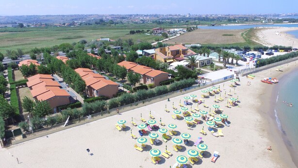 Una località balneare con diversi edifici in stile bungalow, una spiaggia sabbiosa con numerosi ombrelloni e vista sul mare e sul paesaggio circostante.
