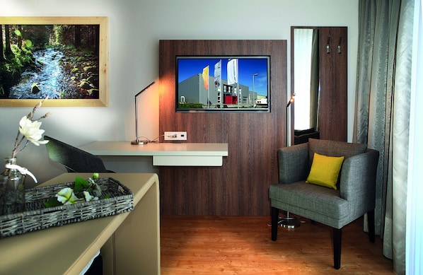 Una habitación moderna con escritorio, un sillón cómodo, televisión montada en una pared de madera y un gran ventanal con cortinas.