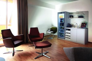 Ein modernes Wohnzimmer mit zwei roten Sesseln, einem grauen Sofa, einem kleinen runden Tisch und einer Minibar inklusive Kühlschrank und Regalen.