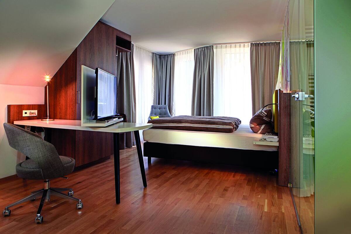 A modern hotel room with a desk, chair, and a bed with a view of the city.