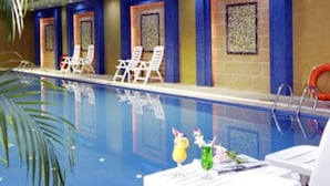 Una piscina interior con agua azul cristalina, sillas de descanso blancas y una pared de azulejos con un diseño decorativo.